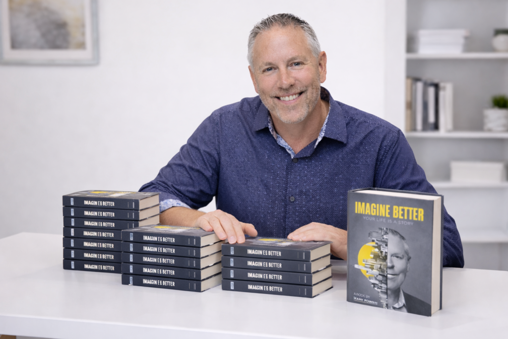 Mark Powers sits behind a clean white desk, smiling confidently with stacks of his book Imagine Better: Your Life Is a Story arranged in front of him, creating a professional and welcoming author portrait.